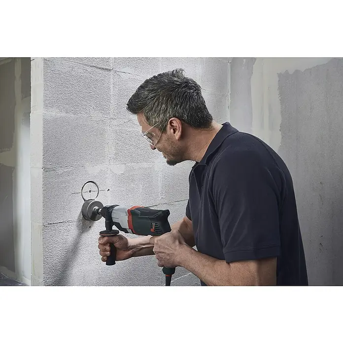 Wolfcraft Plantilla de perforación - Específico para: Instalación de cajetines eléctricos en ladrillo - Un hombre usa una perforadora de núcleo para hacer un agujero en una pared de concreto, con gafas de seguridad.