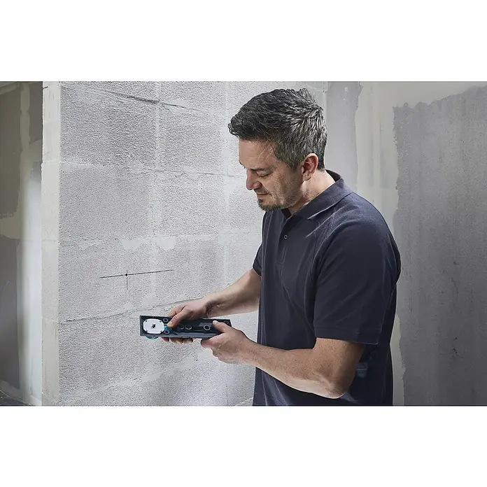 Wolfcraft Plantilla de perforación - Específico para: Instalación de cajetines eléctricos en ladrillo - Un hombre instala un aparato en una pared de ladrillos. La herramienta es gris oscuro con detalles verdes. Centrado en la precisión.