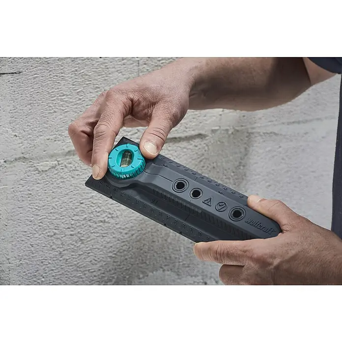Wolfcraft Plantilla de perforación - Específico para: Instalación de cajetines eléctricos en ladrillo - El nivel de burbuja multifuncional Wolfcraft en uso, sostenido por una mano frente a una pared gris.