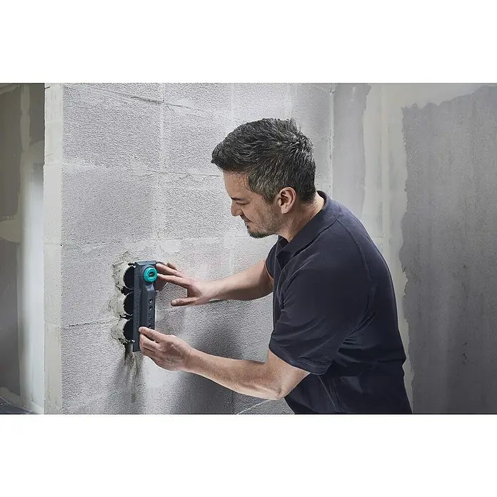 Wolfcraft Plantilla de perforación - Específico para: Instalación de cajetines eléctricos en ladrillo - Un hombre sujeta una herramienta con tres agujeros a una pared. La herramienta es de color gris oscuro con un botón giratorio turquesa.