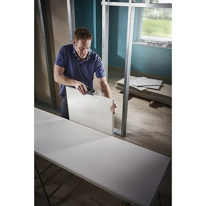 Wolfcraft Escofina plana 4039000 - L x An: 150 x 57 mm, Acero - Un hombre corta una placa de yeso con una herramienta en un banco de trabajo para ajustarla a una pared metálica.