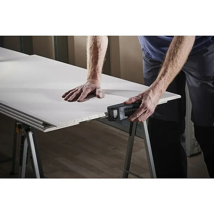 Wolfcraft Escofina plana 4039000 - L x An: 150 x 57 mm, Acero - Un hombre está suavizando una placa de yeso laminado con una herramienta de lijado en un banco de trabajo. La herramienta es negra y tiene un diseño perforado.