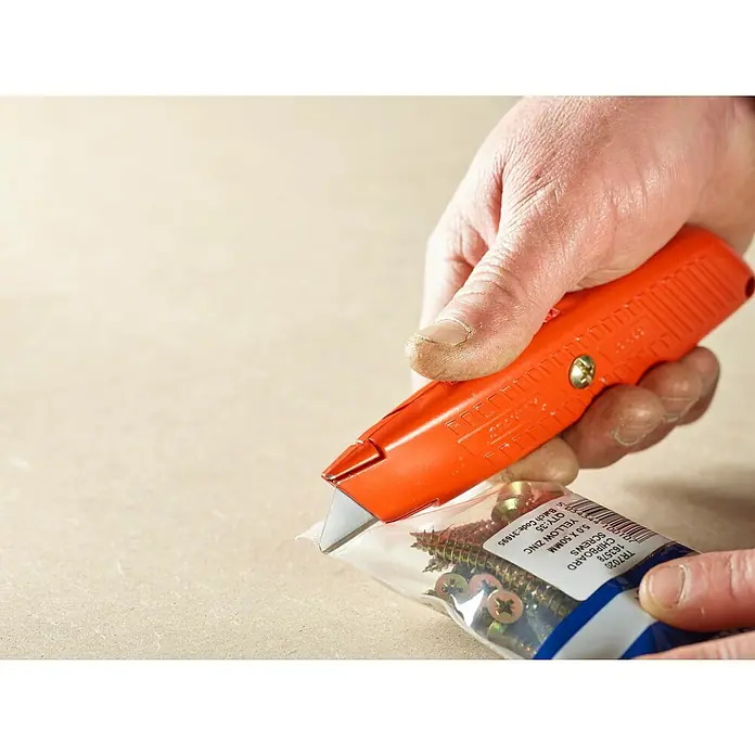 Stanley Cuchillo cúter Tipo 99 E - Largo: 155 mm - Una mano está abriendo un paquete de tornillos para aglomerado amarillos de 5,0 x 50 mm con un cúter naranja.
