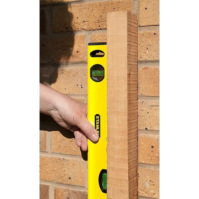 Stanley Nivel de burbuja STHT1-43103 - Aluminio, Amarillo, 60 cm - Se está utilizando un nivel Stanley amarillo para alinear una pieza de madera contra una pared de ladrillos. El nivel indica la verticalidad de la madera.