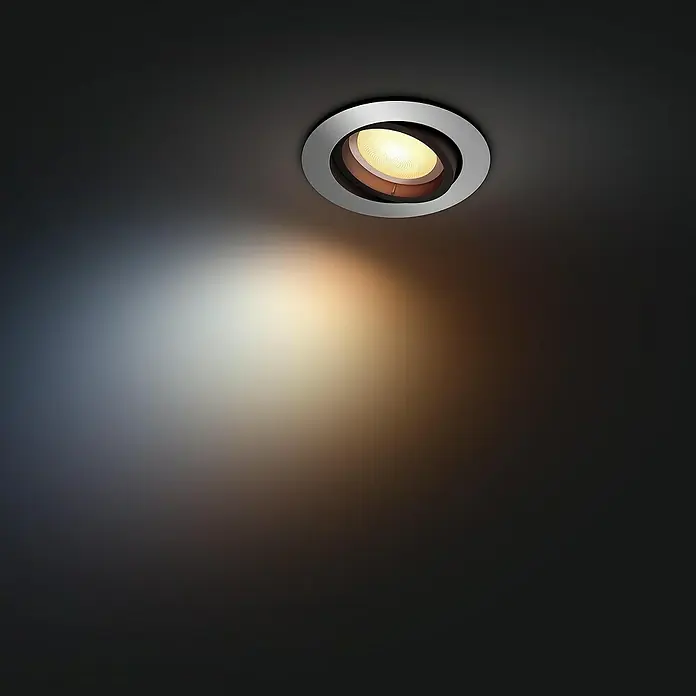 Philips Hue LED-Einbauspot White Ambience Recessed Milliskin - 4,2 W, Ø x H: 9 x 10 cm, Alu, 1 Stk., Kaltweiß - Ein moderner Deckenstrahler mit silberner Einfassung beleuchtet einen dunklen Raum. Das Licht erzeugt einen warmen, einladenden Schein.