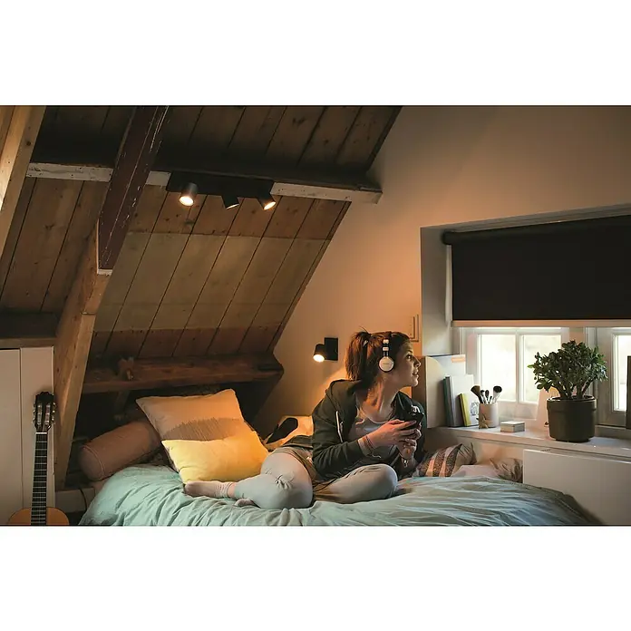 Philips Hue LED-Deckenstrahler White Ambience Runner Erweiterung - 4,2 W, L x B x H: 9 x 11 x 10,7 cm, Schwarz, Mehrfarbig - Junge Frau mit Kopfhörern und Smartphone auf einem Bett in einem Dachzimmer mit Philips Leuchten.