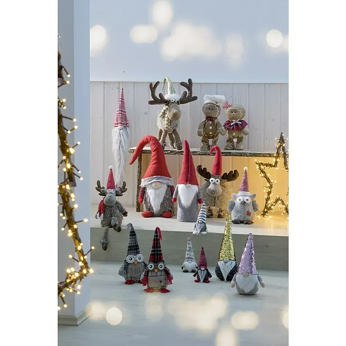 Weihnachtszwerg Weihnachtsmann mit langer Mütze - Textil, Weiß/Rot, Höhe: 52 cm - Eine festliche Anordnung von Weihnachtszwergen und -figuren, die auf einem Tisch und dem Boden arrangiert sind, mit warmen Lichtern im Vordergrund.