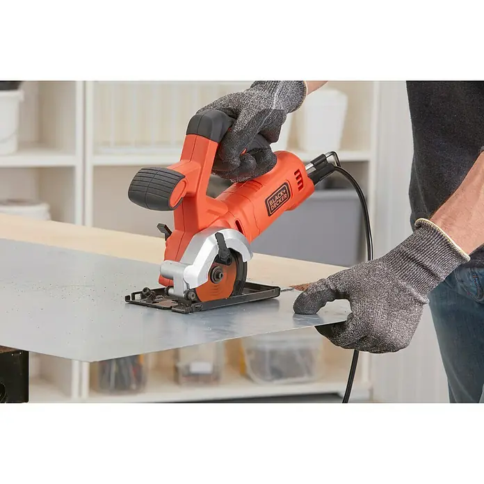 Black+Decker Minisierra circular de mano BES510-QS - 400 W, Diámetro hoja de sierra: 85 mm, Número de revoluciones de marcha en vacío: 3.600 r.p.m. - Un primer plano de una sierra Black+Decker en acción, cortando una lámina metálica, destacando los guantes de protección.