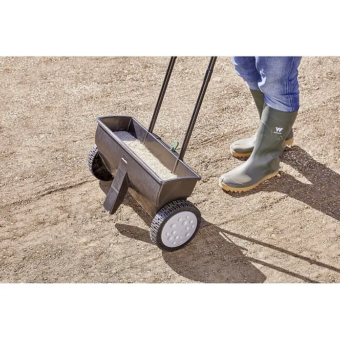 Gardol Carro esparcidor ST 451 - 12 l, Plástico - Imagen de un esparcidor de césped, lleno de semillas, listo para sembrar. Está sobre suelo preparado junto a botas de goma verdes.