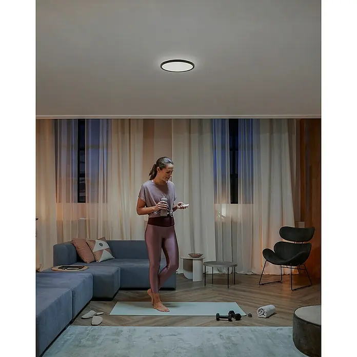 Philips Hue LED-Panel rund White Ambiance Tento S - 16,6 W, Ø x H: 29,1 x 3,2 cm, Schwarz, Kaltweiß - Szene eines Wohnzimmers mit einer schlanken, runden Deckenleuchte, die modernes Design und sanftes Licht vereint.