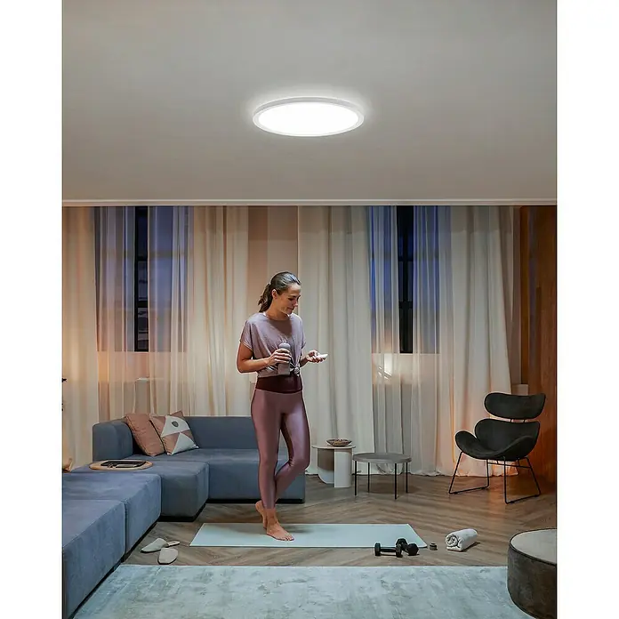 Philips Hue LED-Panel rund White Ambiance Tento XL - 25 W, Ø x H: 54,2 x 3,56 cm, Weiß, Kaltweiß - Die LED-Deckenleuchte erhellt einen modernen Wohnraum mit einer Frau auf einer Yogamatte. Ideal für stilvolle und energieeffiziente Beleuchtung.