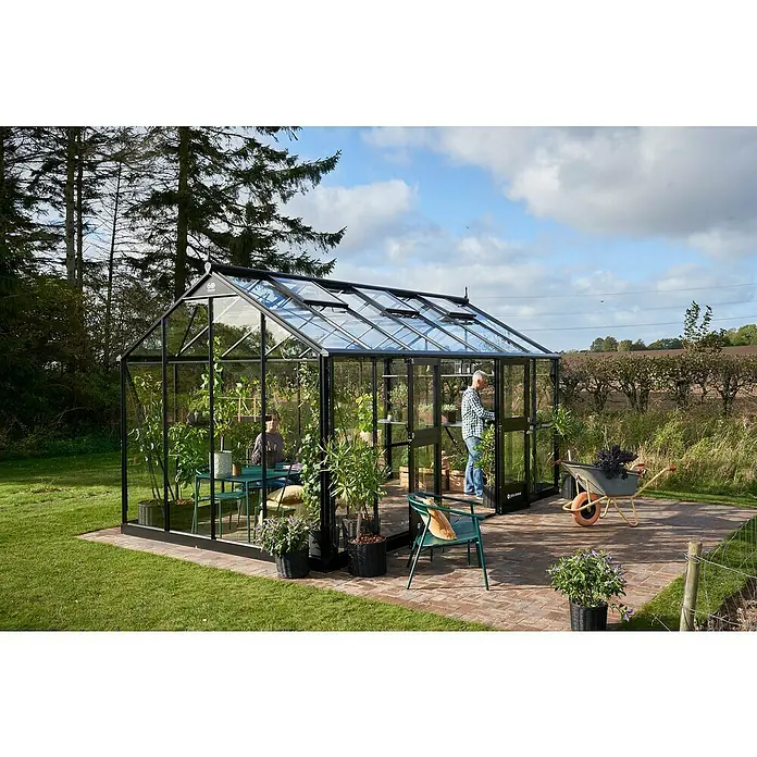 Juliana Gewächshaus Jubii 60 - Material Seitenwand: Glas, 3 mm, Schwarz - Ein modernes Gewächshaus mit schwarzem Rahmen und Glaswänden, umgeben von einem gepflegten Garten mit Bäumen im Hintergrund.