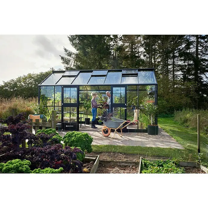 Juliana Gewächshaus Jubii 60 - Material Seitenwand: Glas, 3 mm, Schwarz - Ein Juliana Gewächshaus mit zwei Personen, die Pflanzen austauschen, umgeben von einem Garten mit Gemüsebeeten.