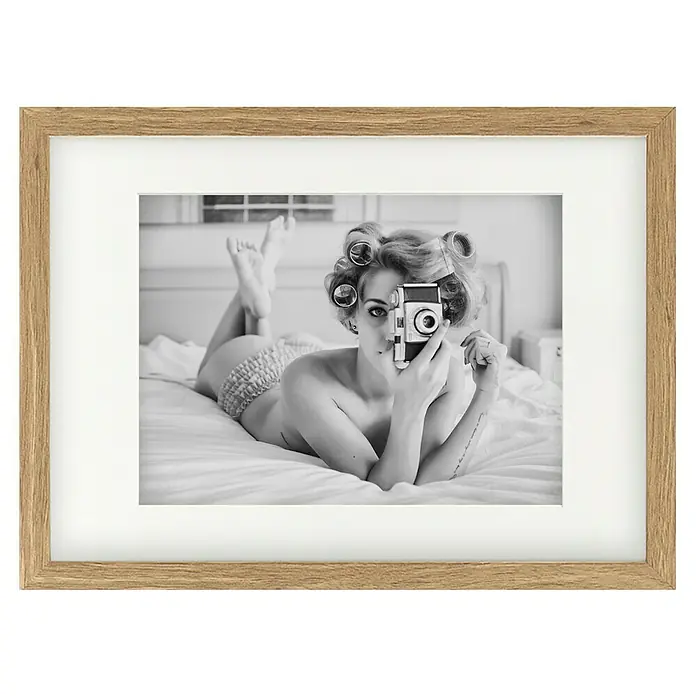 Papermoon Bild - A Chick, B x H: 80 x 60 cm, Mit Passepartout, Rahmen Eiche - Gerahmtes Schwarz-Weiß-Foto einer Frau mit Lockenwicklern, die auf einem Bett liegt und eine Vintage-Kamera hält.