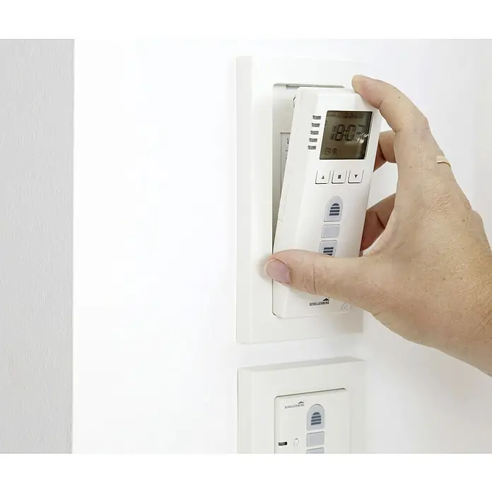 Schellenberg Temporizador inalámbrico Premium - Sistemas soportados: Sistema Smart Home Schellenberg, Apto para: 25 motores telecontrolados - Una mano está colocando un temporizador inalámbrico Schellenberg en un soporte de pared blanco. El dispositivo muestra la hora 18:03.