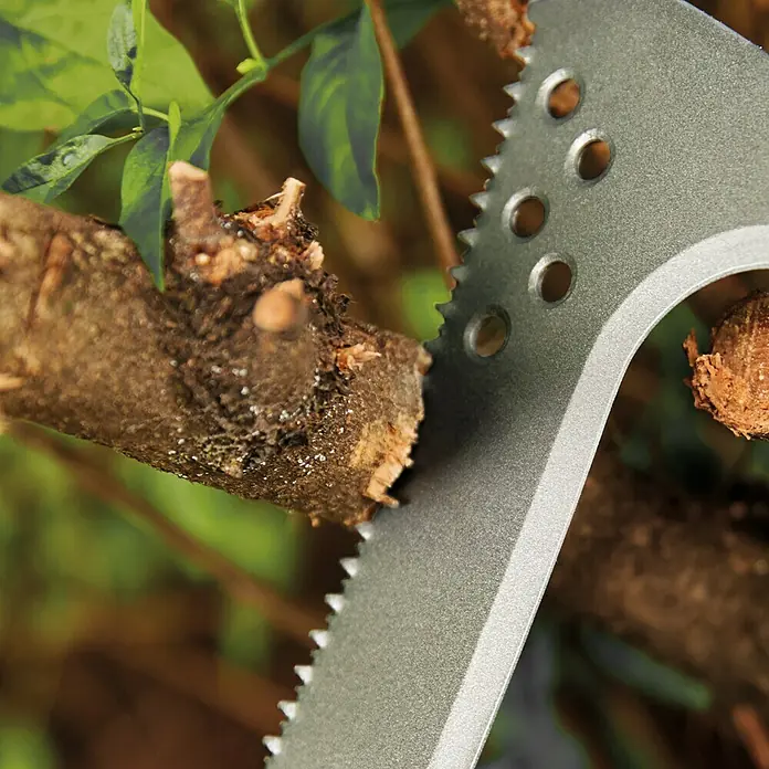 Fiskars Solid Machete mit Sägekante - Länge: 45 cm - Nahaufnahme eines silbernen Multifunktionswerkzeugs, das einen Ast durchtrennt, mit grünen Blättern im Hintergrund.