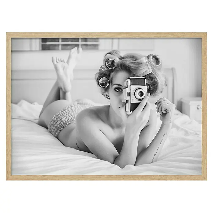 Papermoon Bild - A Chick, B x H: 70 x 50 cm, Ohne Passepartout, Rahmen Eiche - Schwarz-Weiß-Foto einer Frau mit Lockenwicklern und Kamera im Bett, gerahmt in Holz.