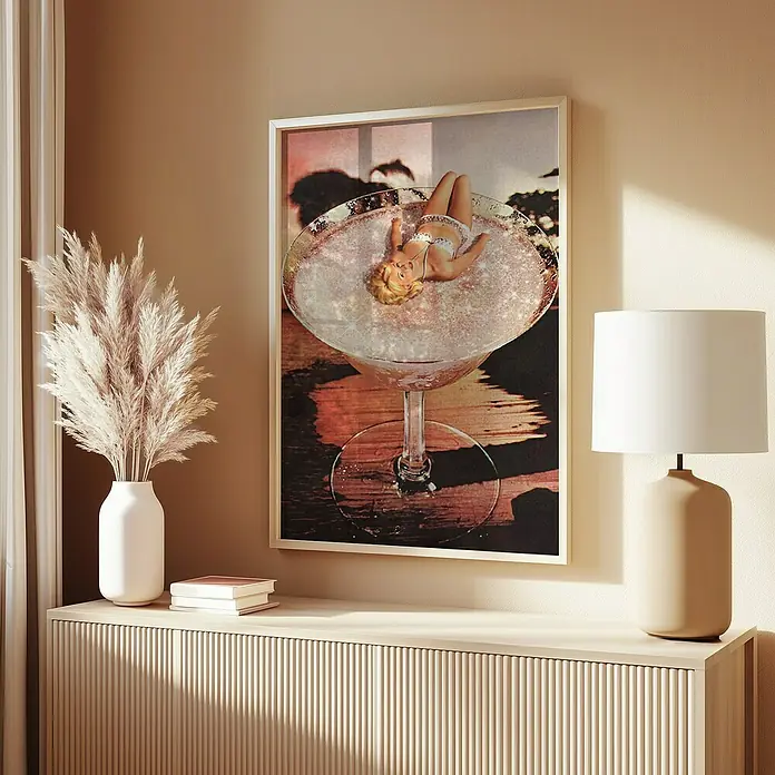 Papermoon Bild - Pink Margarita, B x H: 70 x 100 cm, Ohne Passepartout, Rahmen Eiche - Bild eines gerahmten Kunstdrucks mit einer Frau in einem Martini-Glas, platziert auf einem Sideboard mit Dekor wie einer Lampe und einer Vase.
