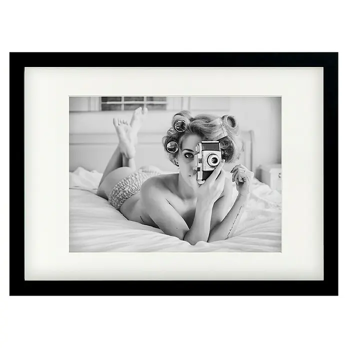 Papermoon Bild - A Chick, B x H: 80 x 60 cm, Mit Passepartout, Rahmen Schwarz - Gerahmtes Schwarz-Weiß-Foto einer Frau mit Lockenwicklern, die auf einem Bett liegt und eine Vintage-Kamera hält, stilvolles Dekor für jeden Raum.
