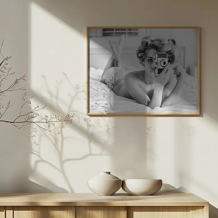 Papermoon Bild - A Chick, B x H: 70 x 50 cm, Ohne Passepartout, Rahmen Eiche - Schwarz-Weiß-Fotografie einer Frau mit Lockenwicklern, die eine Kamera hält und auf einem Bett liegt, gerahmt und an einer Wand befestigt.