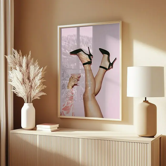 Papermoon Bild - Cheers Peaches, B x H: 80 x 110 cm, Ohne Passepartout, Rahmen Eiche - Das gerahmte Wandbild zeigt Beine mit schwarzen High Heels und einer Hand mit einem Glas vor einem rosa Hintergrund, ergänzt durch eine moderne Kommode mit Lampe.