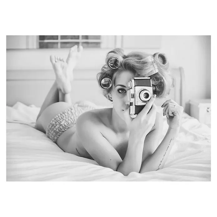 Papermoon Poster - A Chick, B x H: 70 x 50 cm, Ohne Passepartout - Porträt einer Frau mit Lockenwicklern, die auf einem Bett liegt und eine Kamera hält, ein Vintage-Fotografie-Stil, der die Aufmerksamkeit auf Retro-Ästhetik lenkt.