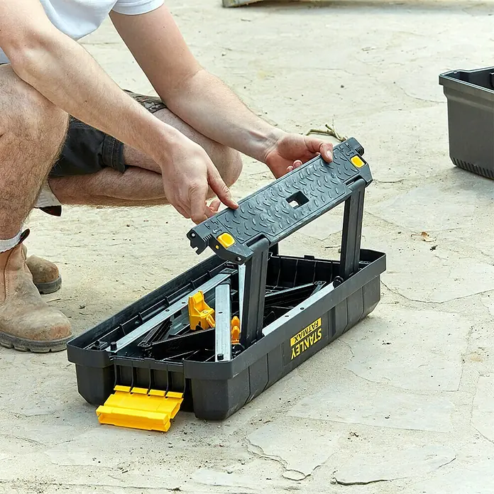 Stanley FatMax Werkzeugkasten FMST81083-1 - L x B x H: 29,6 x 64 x 28,7 cm, Klappbar - Eine Person hockt vor einer Stanley Fatmax Werkzeugkiste und hält ein Werkzeugteil in der Hand.