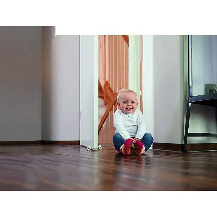 Abus Türstopper Leif - Kunststoff - Glückliches Baby sitzt auf dem Boden vor einer offenen Tür. Das Kind lächelt und trägt bunte Socken.