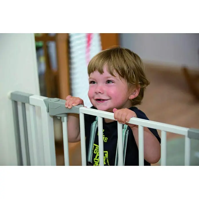 Abus Sicherheitsgitter Finn - Verstellbar: 73 cm - 81,5 cm - Lächelnder Junge, der sich an einem weißen Treppenschutzgitter festhält. Das Gitter ist grau akzentuiert und sorgt für Babysicherheit.