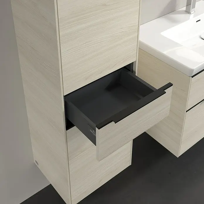 Villeroy & Boch Subway 3.0 Hochschrank - B x T x H: 40 x 36,2 x 171 cm, 1x Schublade, 2x Tür, White Oak, Farbe Griff: Volcano black, DIN Anschlag: Links - Offene Schublade eines modernen Waschtischunterschranks in Holzoptik. Der Waschtisch ist weiß und mit einer Armatur versehen.