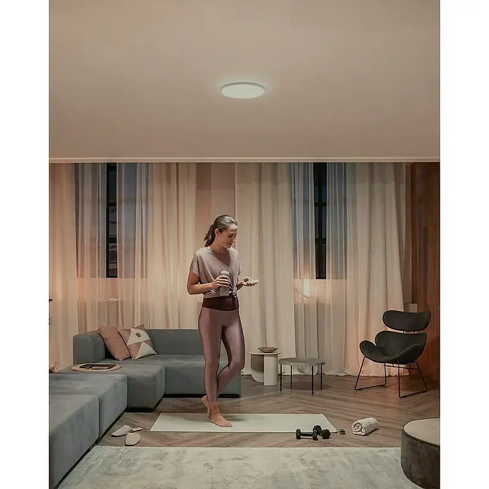 Philips Hue LED-Panel rund White Tento S - 16,6 W, Ø x H: 29,1 x 3,2 cm, Weiß, Warmweiß - Frau mit Getränk neben einer schlanken, runden Deckenleuchte, die ein helles, gleichmäßiges Licht abgibt.