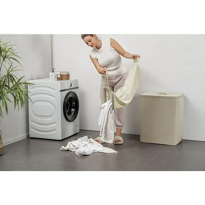 Store It Premium Wäschesammler Ava - 67 x 56 x 40 cm, Beige - Eine Frau leert einen zusammenklappbaren Wäschekorb neben einer Waschmaschine und einem Wäschebehälter, was die einfache Handhabung hervorhebt.
