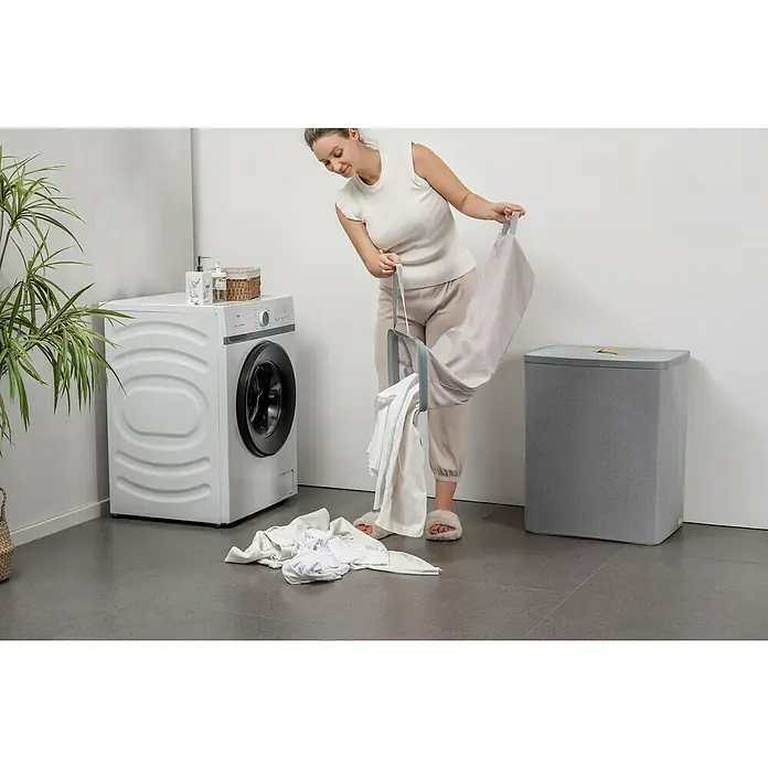 Store It Premium Wäschesammler Ava - 67 x 56 x 40 cm, Grau - Frau mit einem tragbaren Wäschekorb beim Wäschewaschen in einem hellen Raum mit Waschmaschine und Wäschekorb.