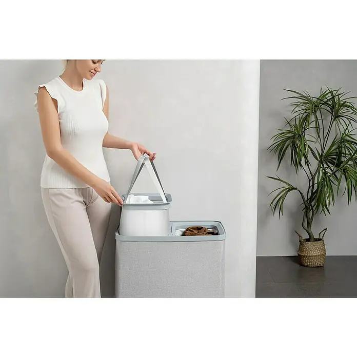 Store It Premium Wäschesammler Ava - 67 x 56 x 40 cm, Grau - Frau, die einen grauen Wäschekorb mit zwei Fächern verwendet. Der Korb verfügt über einen herausnehmbaren Einsatz für einfaches Entleeren.