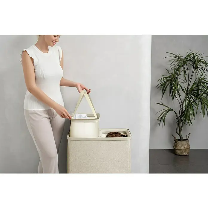 Store It Premium Wäschesammler Ava - 67 x 56 x 40 cm, Beige - Eine Frau, die einen Wäschekorb mit Griffen in einen Wäschebehälter mit zwei Fächern stellt. Praktische Wäscheaufbewahrung.
