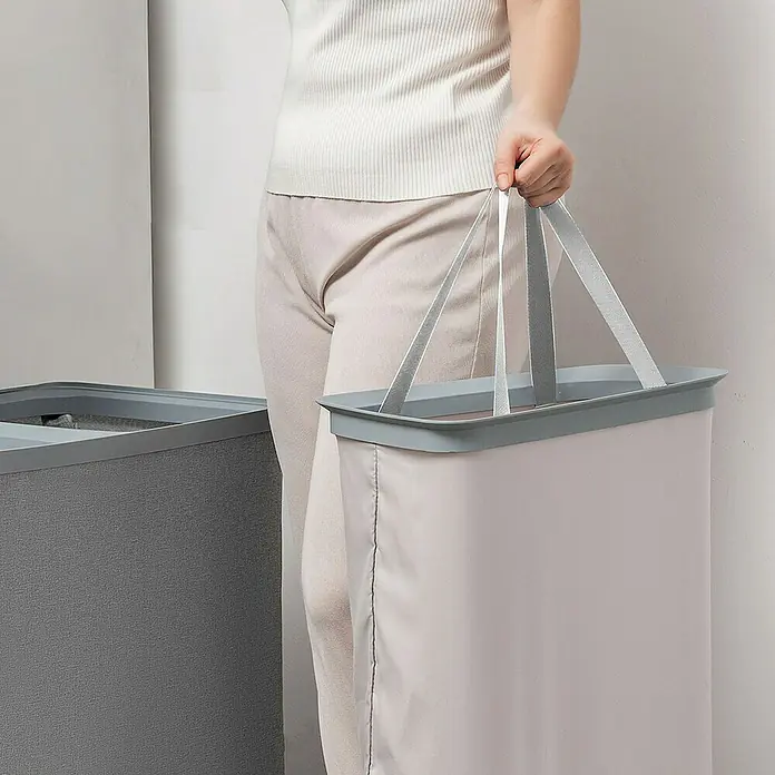 Store It Premium Wäschesammler Ava - 67 x 56 x 40 cm, Grau - Eine Person trägt einen hellen Wäschesammler mit Griffen. Daneben steht ein dunklerer Wäschesammler.