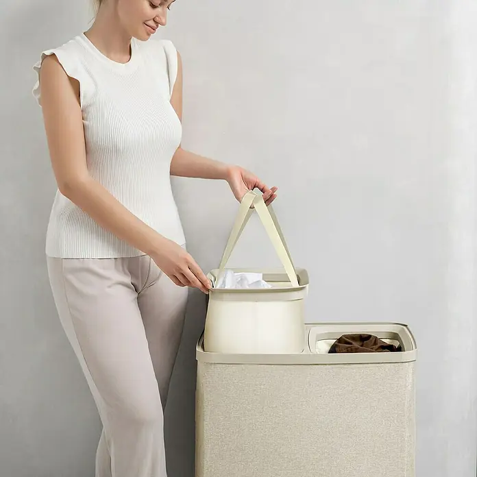 Store It Premium Wäschesammler Ava - 67 x 56 x 40 cm, Beige - Eine Frau befüllt einen Wäschekorb mit zwei Fächern. Der Wäschekorb erleichtert das Sortieren der Wäsche vor dem Waschen.