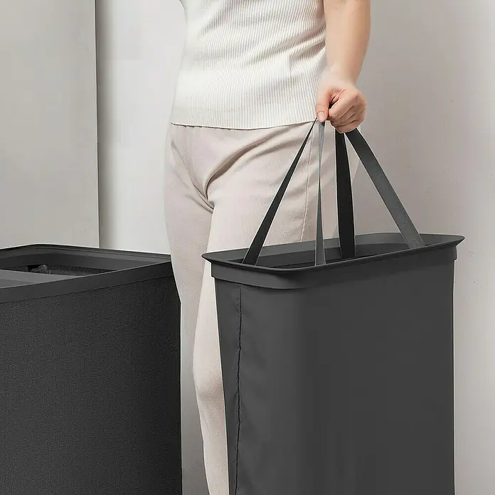 Store It Premium Wäschesammler Ava - 67 x 56 x 40 cm, Schwarz - Eine Person hält einen grauen Wäschekorb mit Griffen. Der Korb ist rechteckig und steht neben einem ähnlichen Wäschekorb.