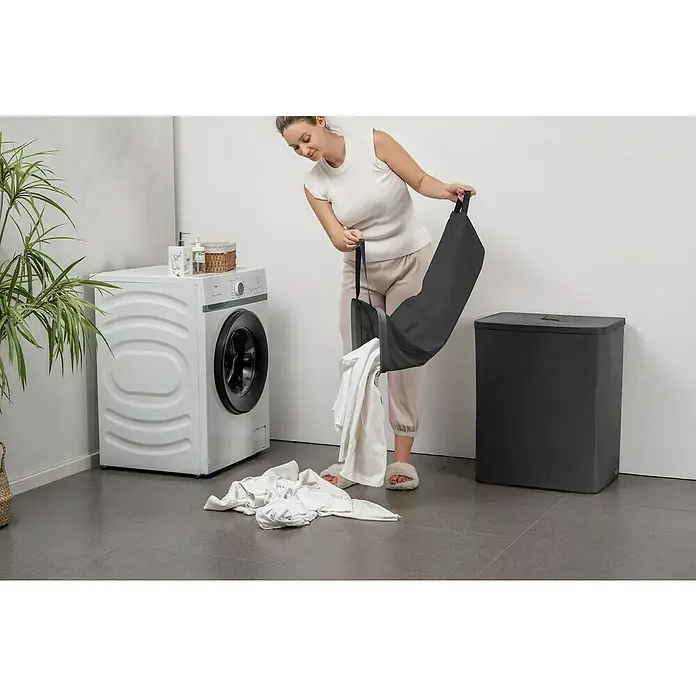 Store It Premium Wäschesammler Ava - 67 x 56 x 40 cm, Schwarz - Eine Frau leert eine graue Wäschetasche, wobei weiße Wäsche auf dem Boden landet, im Hintergrund Waschmaschine und Wäschekorb.