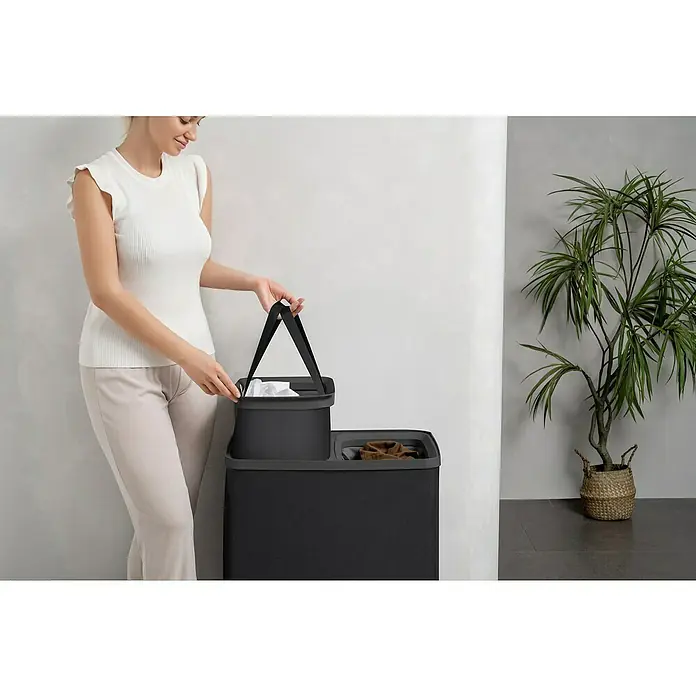 Store It Premium Wäschesammler Ava - 67 x 56 x 40 cm, Schwarz - Frau mit einem schwarzen Wäschekorb mit herausnehmbaren Taschen für die Wäschesortierung und einem Pflanzgefäß im Hintergrund.