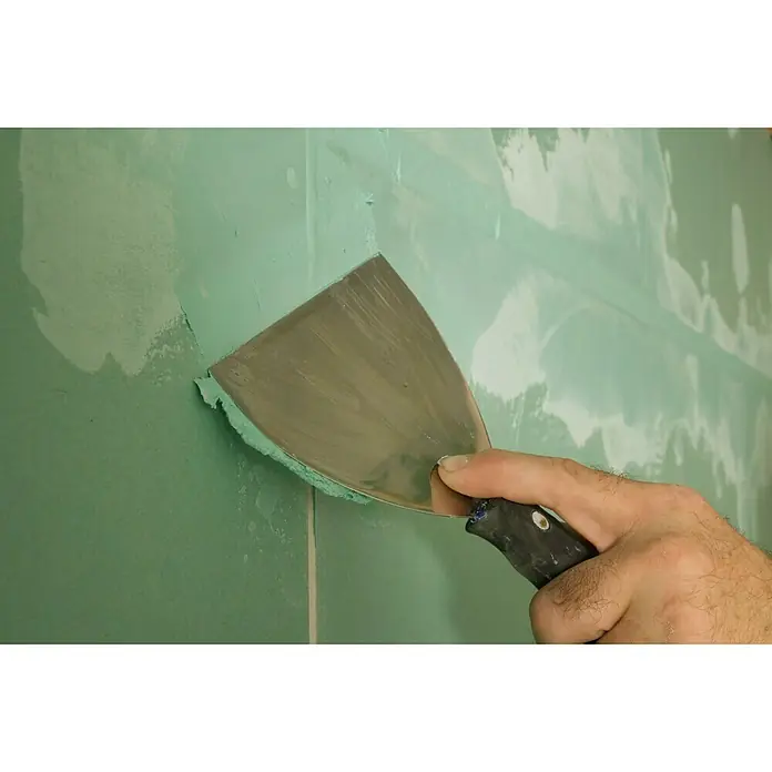 Knauf Fugenspachtel Uniflott imprägniert - 5 kg - Ein Handwerker glättet Spachtelmasse an einer grünen Wand mit einem Spachtel. Das Werkzeug ist aus Metall mit schwarzem Griff.