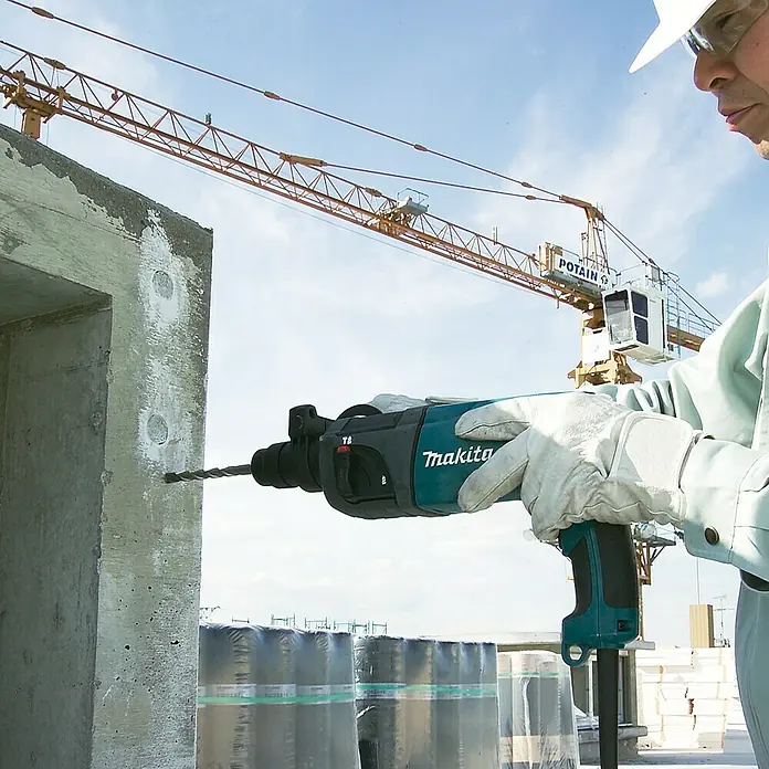 Makita Martillo perforador HR2230 - 710 W, Alojamiento SDS-Plus - Un trabajador de la construcción utiliza un martillo rotativo Makita para perforar un agujero en una pared de hormigón, con una grúa de construcción al fondo.