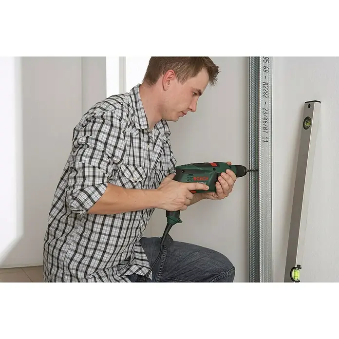 Knauf CW-Standprofil - 3.500 x 100 x 50 mm, Stahl - Ein Mann kniet und bohrt mit einer grünen Bosch-Bohrmaschine in eine Metallschiene an einer weißen Wand.