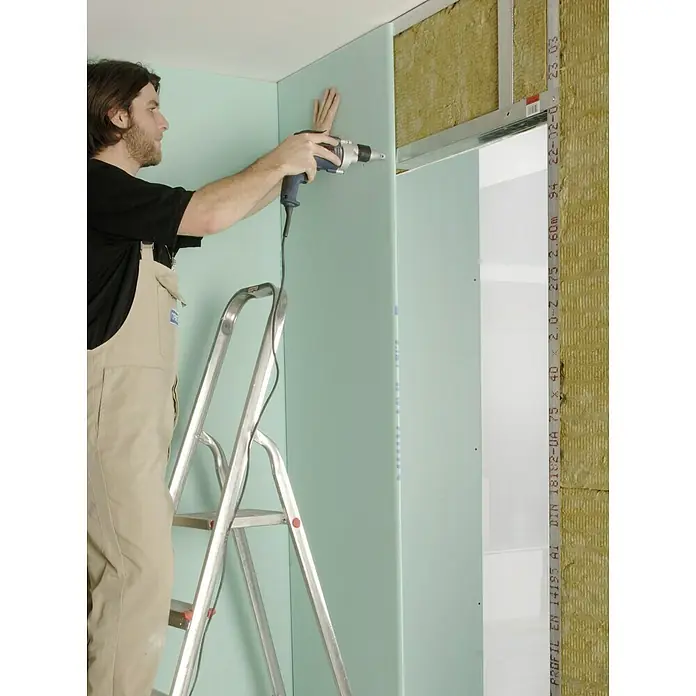 Knauf Schnellbauschraube TN 35 - Durchmesser: 3,5 mm, Länge: 35 mm, 250 Stk. - Ein Handwerker befestigt eine grüne Gipskartonplatte mit einem blauen Akku-Schrauber an einer Wand. Er steht auf einer silbernen Leiter.