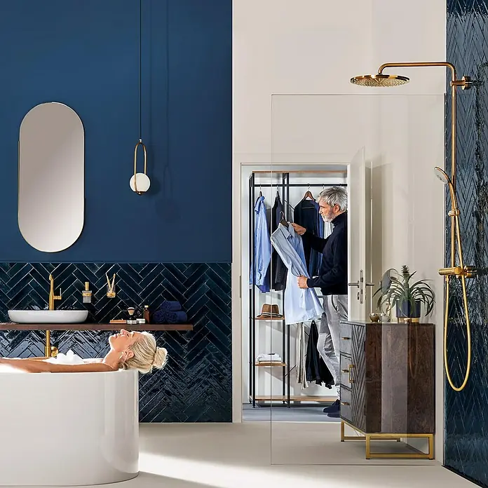 Grohe Essence New Waschtischarmatur XL-Size - Cool Sunrise, Matt - Badezimmer mit Frau in Badewanne, Mann wählt Kleidung aus offenem Schrank. Elegante Armaturen und Accessoires in Gold setzen Akzente.