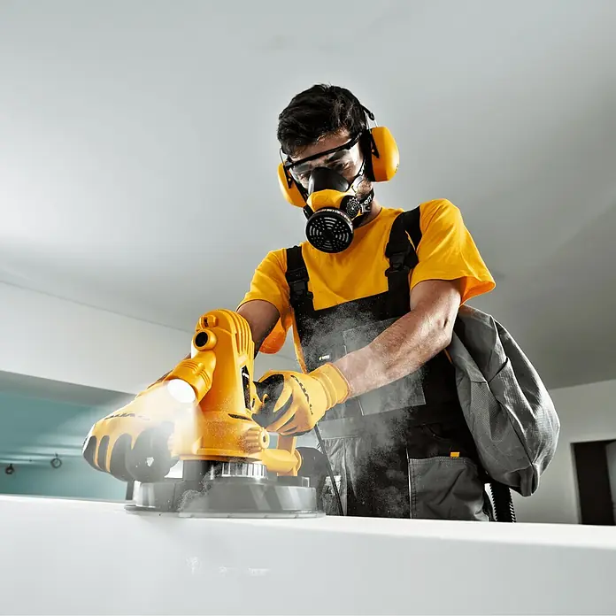 Vito Lijadora de pared y techo VILG800 - 800 W, Diámetro disco de lija: 235 mm - Un artesano con ropa de trabajo amarilla y máscara respiratoria opera una lijadora amarilla, con partículas de polvo visibles en el aire.