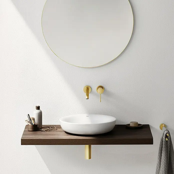 Grohe Essence Waschtischarmatur M - Wandmontage, Cool Sunrise, Matt - Elegantes Badezimmerwaschbecken mit runder Spiegel und Armaturen in Goldoptik. Das Waschbecken steht auf einer dunklen Holzkonsole mit goldenem Standfuß.