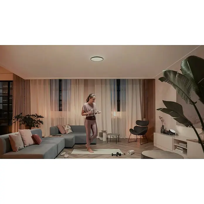 Philips Hue LED-Panel rund White Tento S - 16,6 W, Ø x H: 29,1 x 3,2 cm, Schwarz, Warmweiß - Frau in einem Wohnzimmer mit einer modernen Deckenleuchte. Die Leuchte ist schlicht und elegant.