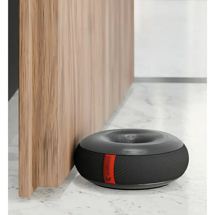 Walteco Türstopper Doornut - Ø x H: 106 x 42 mm, Schwarz - Moderner, runder Türstopper in Grau und Schwarz mit rotem Logo, der eine Holztür schützt und auf einem Marmorboden steht.