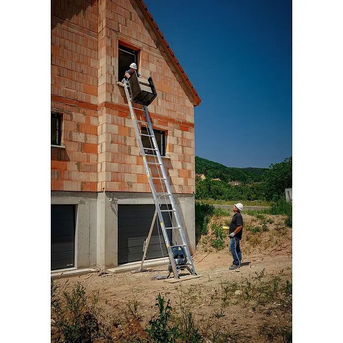 Haemmerlin Maxial Bauaufzug Premium 175/20 Dachdecker - Länge: 10,5 m, Aluminium, Ausführungseigenschaft: Dachdeckerpritsche - Ein Bauarbeiter benutzt einen Schrägaufzug, um Baumaterial in ein Haus zu befördern. Der Schrägaufzug ist zentral im Bild und ein Helfer steht unten.